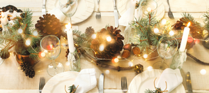 Festive holiday table setting with natural decor, including pine branches, large pinecones, lit candles, and twinkling fairy lights. The table features white plates, glassware, and cloth napkins, creating a warm and rustic ambiance for a holiday gathering.